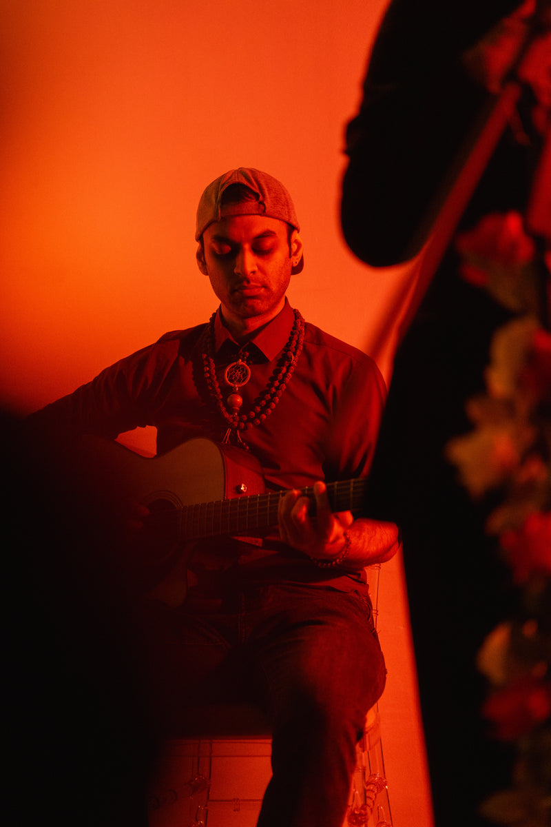 A man wearing a beige baseball cap backwards, a dark red button-down shirt, and layered beaded necklaces sits and plays an acoustic guitar under dramatic orange-red stage lighting.